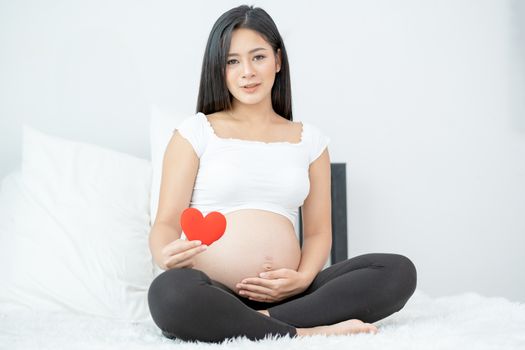 Portrait of beautiful Asian pregnant woman sit on bed and hold red heart icon also smiling and look to camera. Concept of good take care baby in the womb of healthy mother and stay at home.