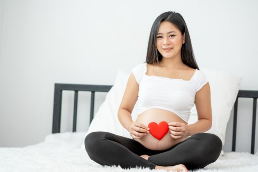 Portrait of beautiful Asian pregnant woman sit on bed and hold red heart icon also smiling and look to camera. Concept of good take care baby in the womb of healthy mother and stay at home.