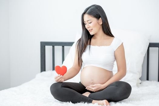 Portrait of beautiful Asian pregnant woman sit on bed and hold red heart icon also smiling and look to symbol. Concept of good take care baby in the womb of healthy mother and stay at home.