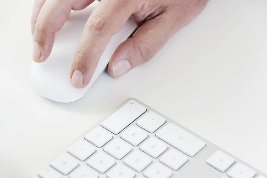 male hand clicking on a white mouse with a white keyboard on the right. White table in a office. Technology and business