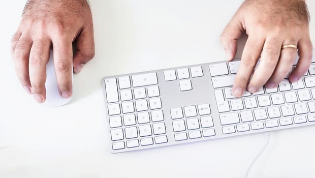 male hand clicking on a mouse and typing on a white keyboard on a white table. White background.
