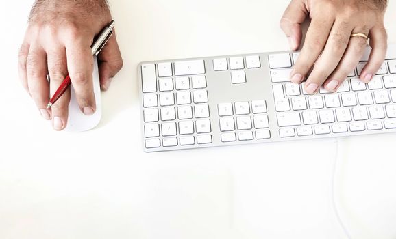 a male while using the computer keyboard and mouse holding out a red pen in his right hand. Technology and creativity. Creative work