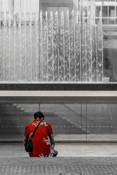 A man in a red shirt sits in front of a fountain, image of street photography with selective desaturation