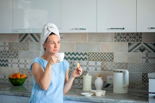 Happy Girl with a towel on her head sutra drinks coffee with cookies for breakfast in his own kitchen