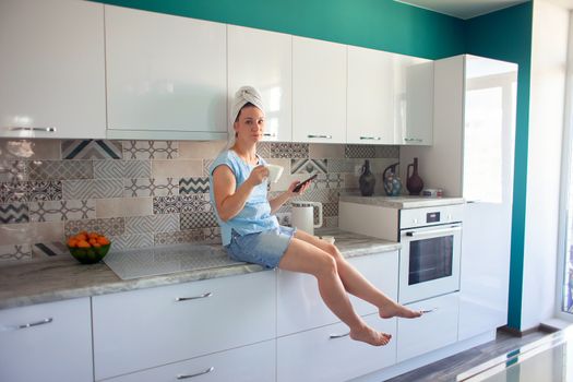 A girl with a towel on her head in her kitchen drinks coffee and looks at the phone