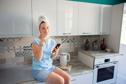 A girl with a towel on her head in her kitchen drinks coffee and looks at the phone
