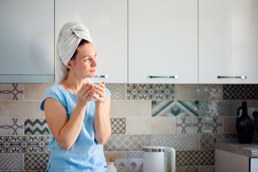 A woman washed her hair in the morning and drinks coffee with a towel on her head. Morning toilet and breakfast in the kitchen