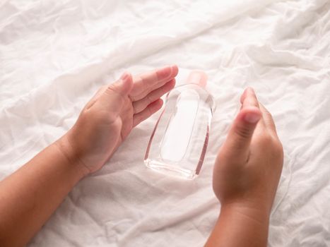 Close-up of Little hands holding transparent plastic bottle of oil on white fabric background. Care about soft body baby skin.