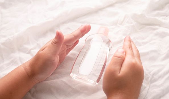 Close-up of Little hands holding transparent plastic bottle of oil on white fabric background. Care about soft body baby skin.