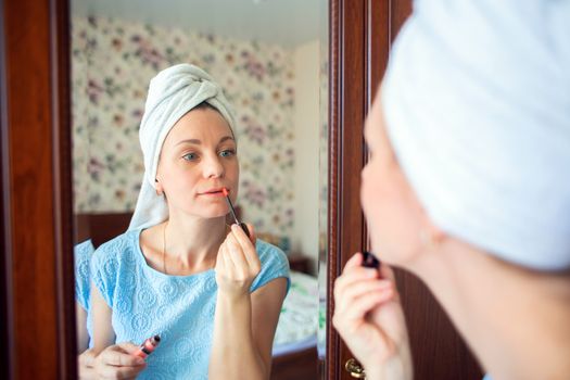 A girl with a towel on her head after a shower paints her lips with lipstick near the mirror. Woman getting ready for work or date