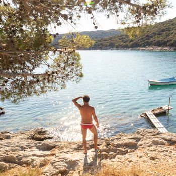 Rear view of man wearing red speedos tanning and realaxing on wild cove of Adriatic sea on a beach in shade of pine tree. Relaxed healthy lifestyle concept.