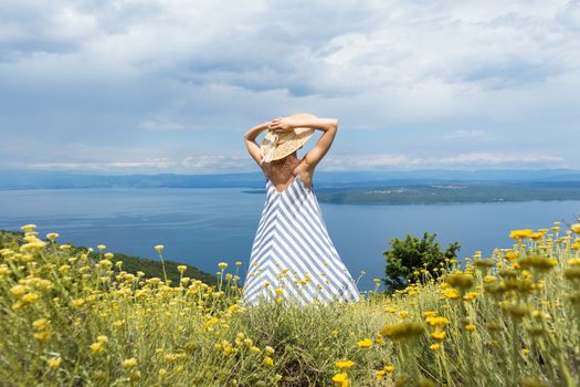 Rear view of young woman wearing striped summer dress and straw hat standing in super bloom of wildflowers, relaxing while enjoing beautiful view of Adriatic sea nature, Croatia.