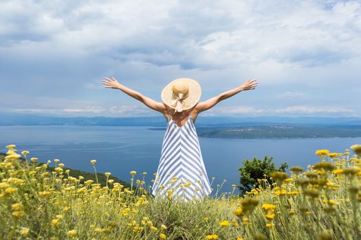 Rear view of young woman wearing striped summer dress and straw hat standing in super bloom of wildflowers, relaxing with hands up to the sky, enjoing beautiful view of Adriatic sea nature, Croatia.