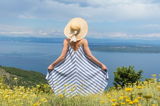 Rear view of young woman wearing striped summer dress and straw hat standing in super bloom of wildflowers, relaxing while enjoing beautiful view of Adriatic sea nature, Croatia.