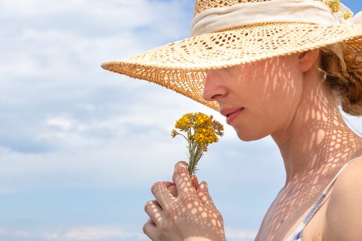 Portrait of young beautiful cheerful woman wearing straw sun hat, smelling small bouquet of yellow wild florets, against blue summer sky.