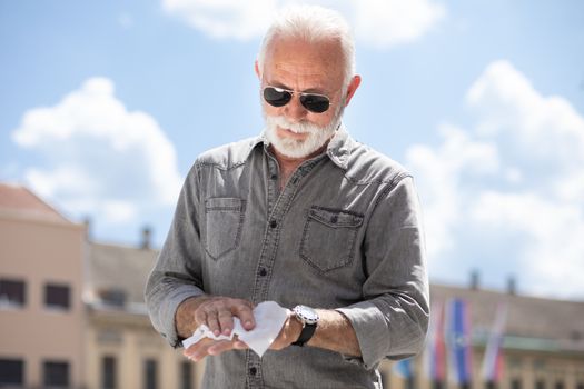 Old man cleaning hands with wet wipes outdoor in public, hot summer day, sunglasses