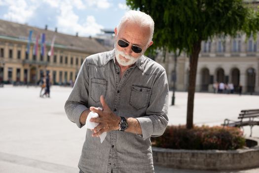 Old man cleaning hands with wet wipes outdoor in public, hot summer day, sunglasses