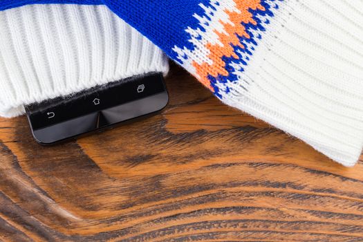 a cellphone inside blue and white knited mittens on wooden background.