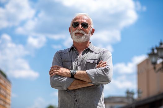Old rich man with beard and sun glasse on street posing and smiling, portrait, blurred clouds background