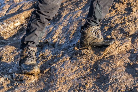 legs in gray pants and trek boots hiking upwards on muddy hill at evening sunlight.