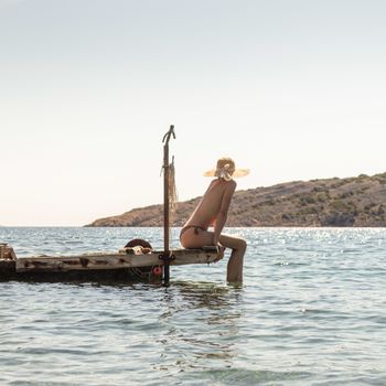 View of unrecognizable woman wearing big summer sun hat tanning topless and relaxing on old wooden pier in remote calm cove of Adriatic sea, Croatia.