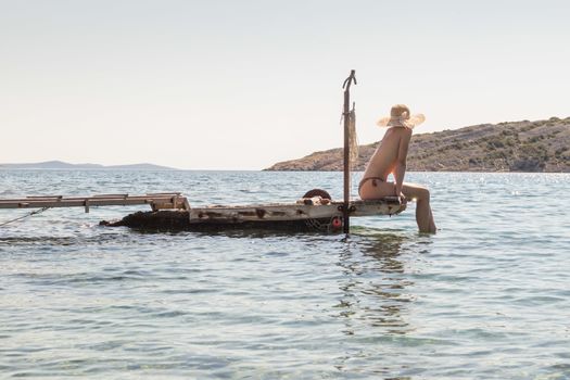 View of unrecognizable woman wearing big summer sun hat tanning topless and relaxing on old wooden pier in remote calm cove of Adriatic sea, Croatia.