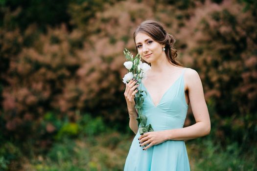 Happy girl in a turquoise long dress in a green park on a background of herbs, trees and rose bushes