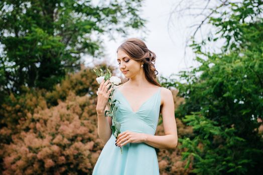 Happy girl in a turquoise long dress in a green park on a background of herbs, trees and rose bushes