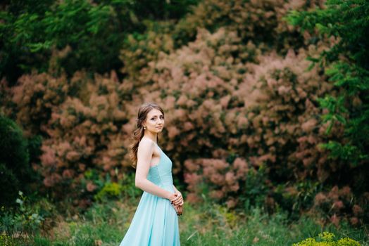 Happy girl in a turquoise long dress in a green park on a background of herbs, trees and rose bushes