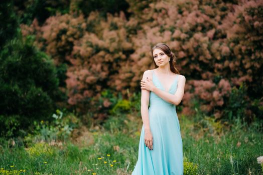 Happy girl in a turquoise long dress in a green park on a background of herbs, trees and rose bushes