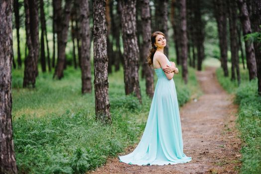 Happy girl in a turquoise long dress in a green park on a background of herbs, trees and rose bushes