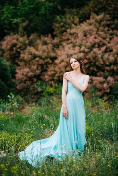 Happy girl in a turquoise long dress in a green park on a background of herbs, trees and rose bushes