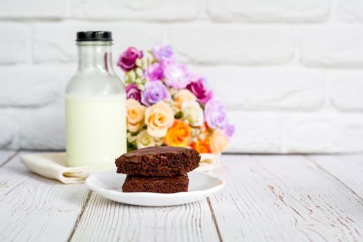 Piece of brownie cake on white small plate with bottle of fresh milk on wooden table and with brick wall with copy space.