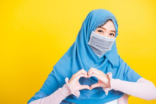 Asian Muslim Arab, Portrait of happy beautiful young woman Islam religious wear veil hijab and face mask protect quarantines disease coronavirus show hands finger heart figure isolated yellow