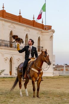 A handsome Mexican Charro poses in front of a hacienda in the Mexican countryside while holding a falcon