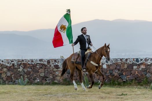 A handsome Mexican Charro poses in front of a hacienda in the Mexican countryside while holding the Mexican Flag