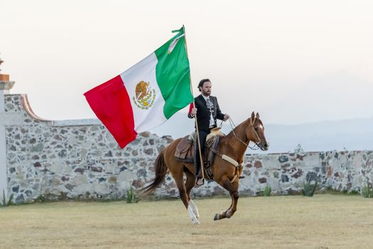 A handsome Mexican Charro poses in front of a hacienda in the Mexican countryside while holding the Mexican Flag