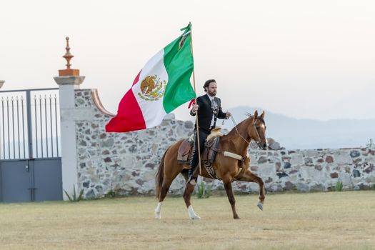 A handsome Mexican Charro poses in front of a hacienda in the Mexican countryside while holding the Mexican Flag