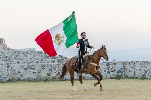 A handsome Mexican Charro poses in front of a hacienda in the Mexican countryside while holding the Mexican Flag