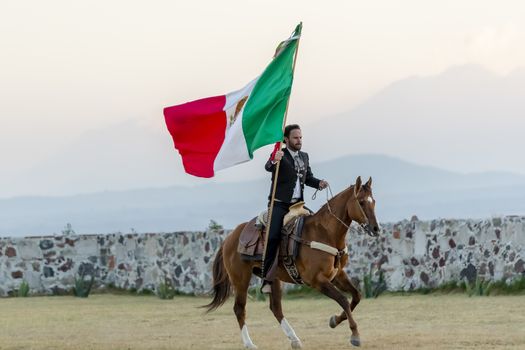 A handsome Mexican Charro poses in front of a hacienda in the Mexican countryside while holding the Mexican Flag