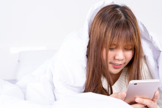 Happy Asian girl using a mobile phone lying under a blanket on the bed at home. Social and technology concept.