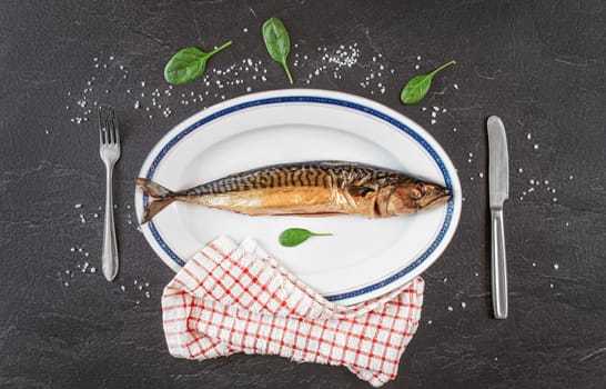 Whole smoked mackerel fish on white oval plate, gray black stone like table under, rock salt and green leaves salad near. View from above.