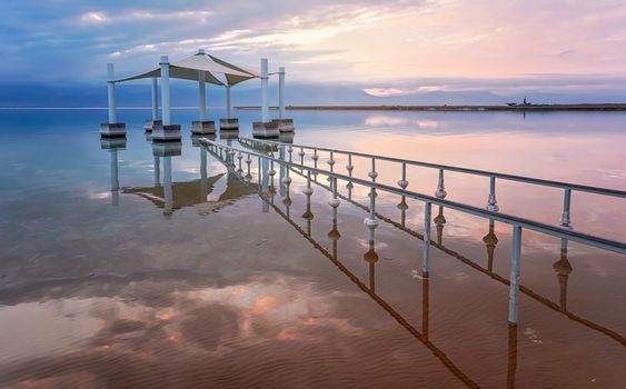Calm morning at Ein Bokek Dead Sea beach, pink clouds reflection over water surface near steel rails leading to sun shade shelter.