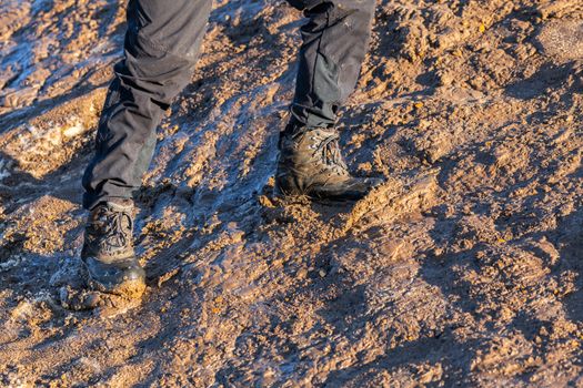 legs in gray pants and trek boots hiking upwards on muddy hill at evening sunlight.