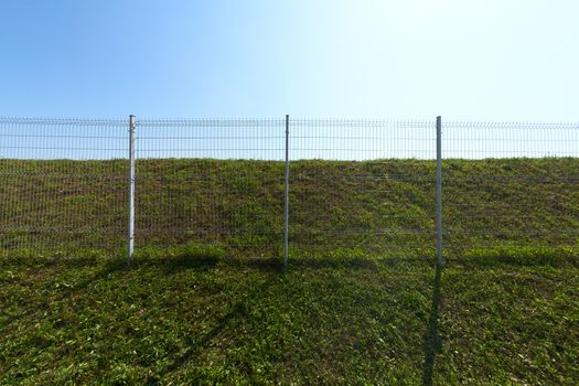 Metal wire industrial grid fence on green grass with blue sky background wide angle horizontal shot.