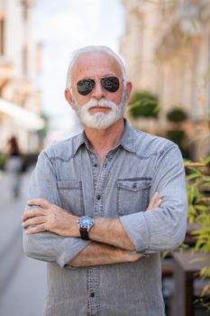 Happy rich old man with beard and sun glasses on street posing and smiling, portrait, blurred city street background