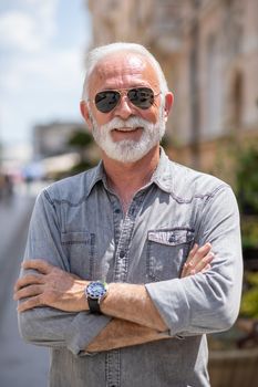 Happy rich old man with beard and sun glasse on street posing and smiling, portrait, blurred city street background