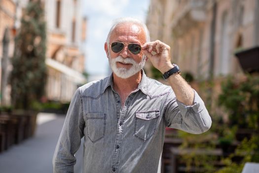 Happy rich old man with beard and sun glasses on street posing and smiling, portrait, blurred city background