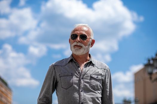 Old man with beard and sun glasses on street posing and smiling, portrait, blurred city street background