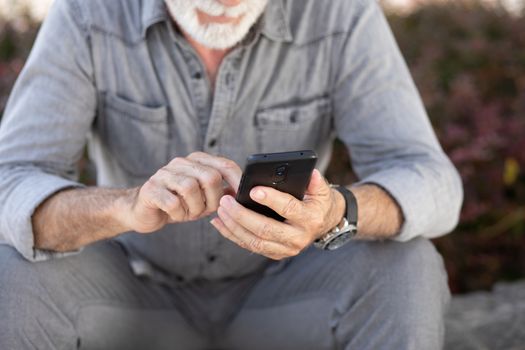 Old man in gray clothing seating on street and typing SMS on smartphone